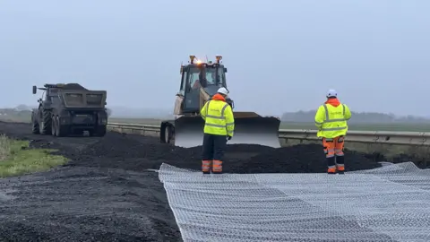 Mousumi Bakshi/BBC A tractor and a digger are on parts of a road which has now got lots of soil on it. There are two people wearing Hi-Vis jackets and are looking at the digger which is scooping some of the soil. A large white sheet is covering part of the road.