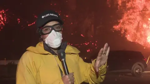 CBS reporter Danya Bacchus wears a face mask and goggles as she reports from the scene of a wildfire in Los Angeles