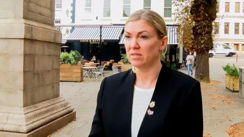 Deputy Raluca Kovacs, a woman with blonde hair which is tied back. She is wearing a black blazer and white top. She has two badges on her lapel. She is standing in Jersey's Royal Square, in front of the entrance to the Cock and Bottle pub. There are orange, autumnal leaves on the ground beneath the trees in the square. 