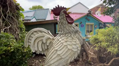 An enormous white cockerel in the garden of a blue, pink and white wooden house