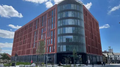 The seven-storey building with red terracotta tiles around the windows on each side and a large rounded glass corner over the entrance that stretches from the first floor to the roof.