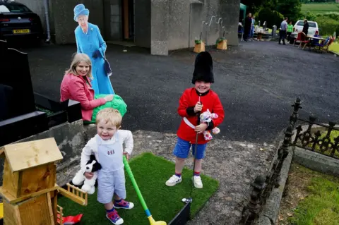 PA Media Laura McKnight holds a cardboard cut-out of Queen Elizabeth II as her sons Noah (5) and Louis (2) play mini golf at a picnic at St Bartholomew's Parish Church, Newry, as part of the Big Jubilee Lunch