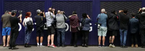 Reuters Photographers and videographers peer into a car park trying to get a glimpse of Serbian tennis player Novak Djokovic at the offices of his legal team