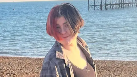 EJ EJ stands on a pebble beach on a sunny day with the shoreline and sea visible in the background. Part of the metal frame of an old pier can also be seen. EJ's hair - which is half-brunette, half-red, partly covers EJ's smiling face. EJ wears a black vest top with an unbuttoned grey/black checked shirt over the top.