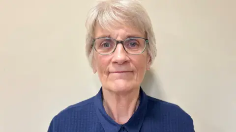 The Cathedral of St John the Baptist Angela Stone standing against a cream-painted wall. She has short silvery hair and is wearing grey-framed glasses and a blue shirt done up at the neck. She is looking towards the camera