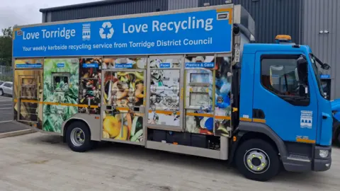 A blue recycling lorry from Torridge District Council. The side of the vehicle has pictures of different types of materials it can recycle including paper, food waste and glass.