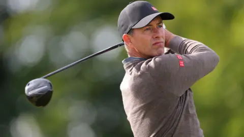 Getty Images Adam Scott watches his golf ball soar into the distance after teeing off with his driver. He is wearing a light brown jumper and a dark grey baseball cap 