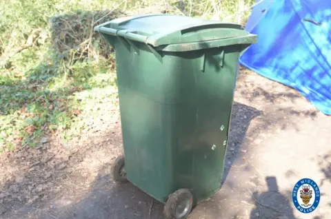 West Midlands Police A green wheelie bin next to a blue police tent behind. It is in the surroundings of a park and greenery can be seen in the background.
