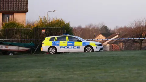 A police car behind a police cordon on a residential street by a patch of grass