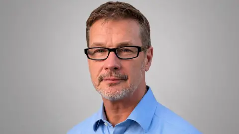 Close-up man with greying hair and greying beard, wearing glasses and a blue shirt