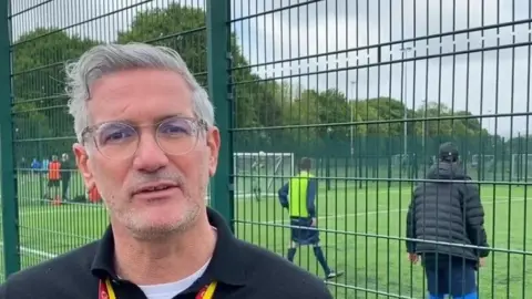 Middlesbrough Council Craig McManus, Acting Head of MFC Foundation, is looking at the camera. He has short grey hair and is wearing a black jumper, yellow and red lanyard and glasses. Players are walking at the football pitch behind him.
