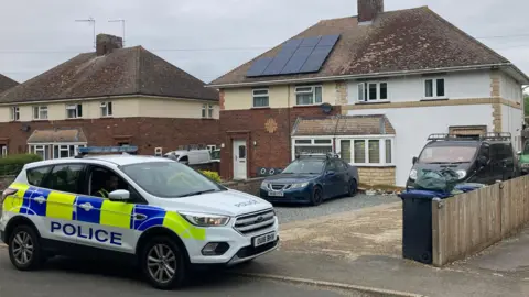 DJMcLaren/BBC A police car outside a semi-detached white house. It is on the left and a car and a van can be seen on the driveway/paved over garden.