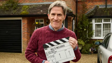 Brydon holding a clapper board with the show's title, outside a house. He wears a red jumper over a blue shirt. He has short grey hair and is smiling at the camera.