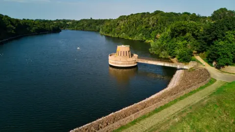There is a large expanse of water in a reservoir with green shrubs and trees beside it and a field and path in front. There is a bridge over the reservoir to a concrete valve tower.