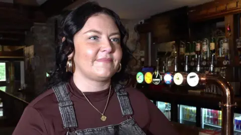 Sophie White stands at the bar of La Ville Hotel in Alderney. She is wearing black dungarees and a maroon top, with a gold medallion. She has black curly hair. Several taps are at the bar including ones for Guiness and Kronenbourg 1664. Bottles of spirit and optics are behind the bar along with fully stocked fridges.