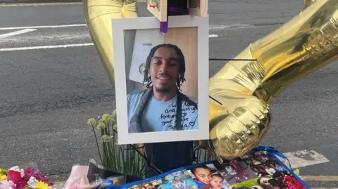 A framed picture of a young man is attached to a lamp post - surrounded by balloons, flowers and other photos of him.