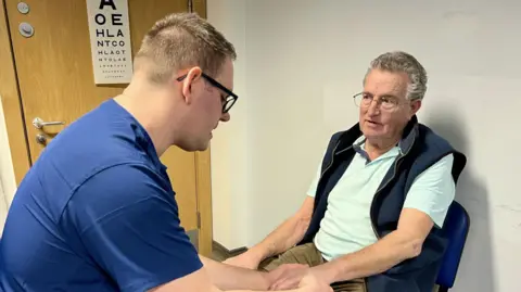 Nikki Fox/BBC Two men in a UTC in Corby. The man on the left has short light brown hair and black-rimmed glasses and is in blue scrubs. He is looking down at the hand of other man, who is sitting on a blue chair, has grey hair and glasses and is wearing a blue gilet over a polo shirt.