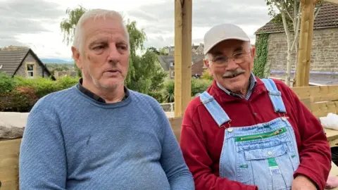 On the left is Steve Hutchings wearing a blue jumper and on the right is Brian Chislett wearing a red jumper and denim dungarees.