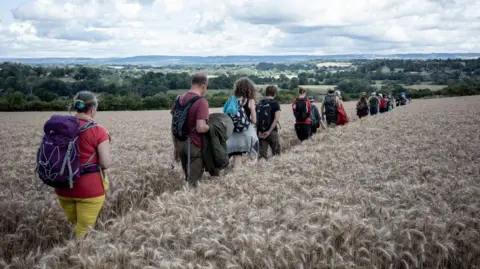 Mark Brumell/Friends of the River Medway A line of people walking through a grain field in a line, travelling away from the camera. A large, green valley can be seen in the distance. The sky is overcast.