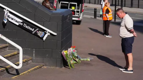 PA Media Lee Dixon standing in front of a row of flowers at the steps of St James' Park. He has grey, short hair and is wearing a beige polo shirt and blue shorts. He has his hands behind his back and his head bowed. There is a Newcastle United scarf attached to the railing to the left.