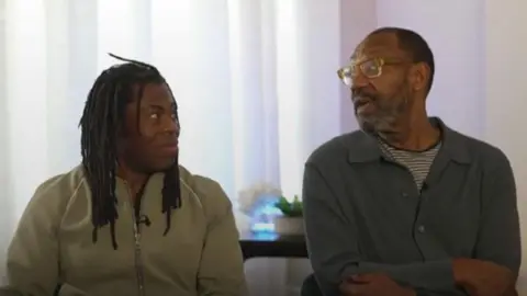 Ade Adepitan, wearing a light green fleece top, is sat next to Sir Lenny Henry, who is wearing glasses and a grey shirt over a striped t-shirt.