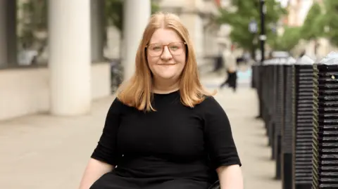Kateryna Pavlyuk A young woman with light shoulder length light and large square-rimmed glasses sits in a wheelchair, smiling at the camera. She wears a black top and skirt, and is outside a large office building on a fair day.