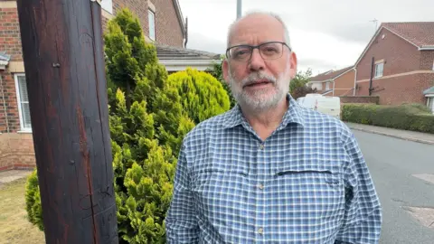 A man wearing glasses and a blue checked shirt stands on a residential street beside a wooden telegraph pole. Brick houses, hedges and parked vehicles are in the background under an overcast sky.