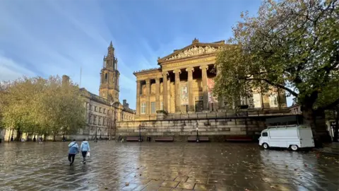 Edwin Williams The site today shows a large paved flat area in front of a church to the left and the facade of the Harris Museum. A large square stone building with pillars at the entrance above a set of steps. Two women are walking past and there is a white van parked to the right under a tree