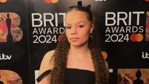 Izzy Withers Izzy Withers on the Brits red carpet. Izzy is a 20-year-old mixed race woman with long brown hair. She wears a black ribbon in her hair and a black off-the-shoulder dress.