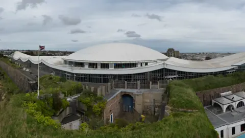 An aerial view of a white dome shaped building with grey skies behind and foliage and brick walls in front.