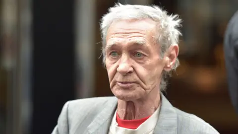 Close up headshot of Paddy Hill looking away from camera while wearing a grey suit against a neutral background, as he attended Council House in Solihull following fresh inquests into the deaths of those in the bombings. Taken in Birmingham in 2016. 