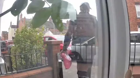 Doorbell camera footage of a man dressed in black with a black cap walking out of a front garden with a pink parcel.