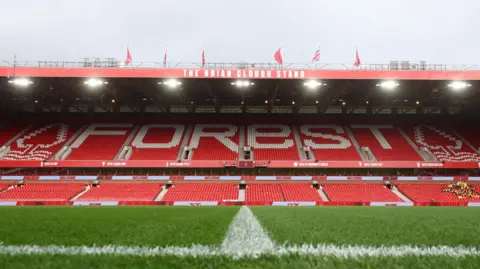A low shot of the Nottingham Forest home pitch, with a tall, red and white football stand in the background.
