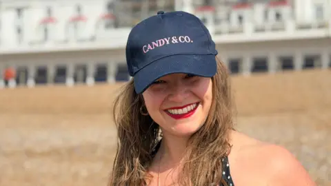 A blonde woman in her 30s wearing a blue baseball cap standing on a beach