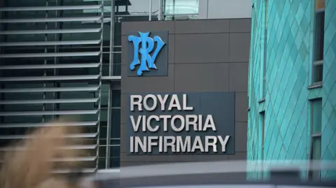 Close up of sign on a hospital which reads in all caps, Royal Victoria Infirmary. There is a blue logo above it made up of the initials RVI. It is on dark cladding next to light blue panelling. The building looks futuristic. 