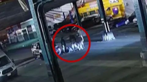 A group of people surround a manhole in the middle of the street. A red circle is around them.