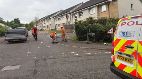 Four workers in high-viz jackets work putting fences up on a residential street next to a parked police van