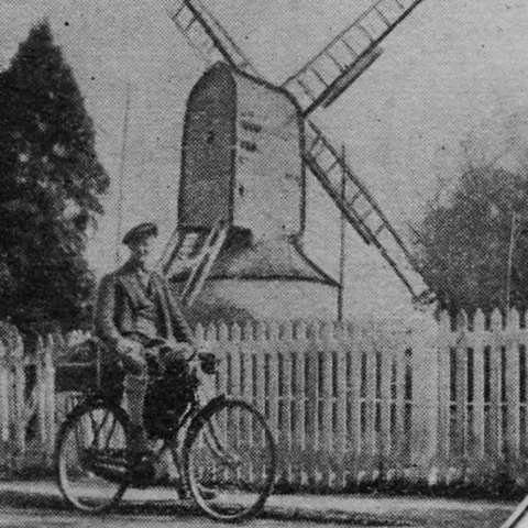 Mills Archive Trust/ C.Burt A grainy black and white picture from a newspaper cutting of a man wearing a beret and riding a bicycle in front of a white fence and wooden windmill