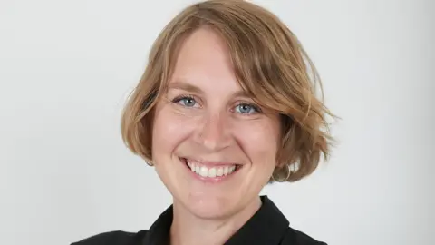British Dyslexia Association Ellen Broomé, CEO of British Dyslexia Association, smiling directly at the camera. She has blonde hair that goes just below her ears. She has blue eyes. She has a silver nose piercing and silver hooped earrings. Behind her is a white wall. She has a black shirt on that has a collar.
