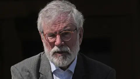 Gerry Adams pictured outside the High Court in Dublin last week wearing a grey suit jacket, a light coloured shirt with the top button undone and a pair of circular brown glasses. Mr Adams has white hair and a beard.
