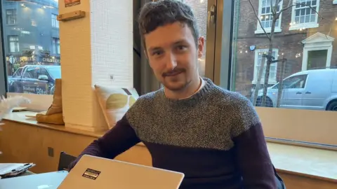 BBC Robert Aston looking into his camera while working on his laptop.