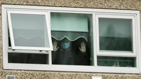 Getty Images A man wearing a mask holds his hands up to the window in one of the housing towers