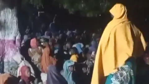 A screengrab from a video showing a crowd of mainly and children, mostly from behind, as they sit under a treat after being freed from Boko Haram captivity. One of the women is standing to the right of the image and wears a yellow headscarf.