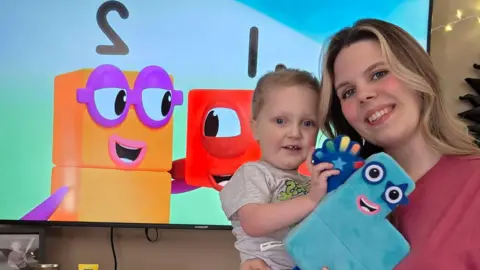 Hannah holding her son Myles, they are smiling at the camera and stood in front of a TV with Numberblocks on. They are also holding Numberblocks toys.
