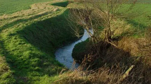 Water flows in a deep ditch with green grass embankment on either side.