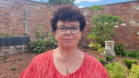 Ellen Knight/BBC Photo of Oliver looking into the camera and slightly smiling. She is wearing a red top and a silver necklace. She is sat in front of a brick wall, which has trees and plants planted along the bottom of it. The sky is blue and there is strong sunshine. 