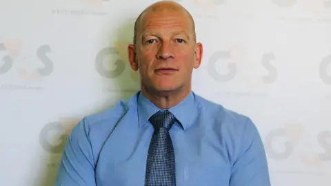 HMP Five Wells Peter Small, looking straight at the camera. He has a bald head, wearing a blue shirt and blue tie. He is standing in front of a wall. He is not smiling. 