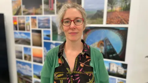 A woman is standing looking directly at the camera. She is wearing a black dress with colourful flowers on it and a green cardigan. She is wearing red glasses. She is standing in front of a wall with photographs on it.