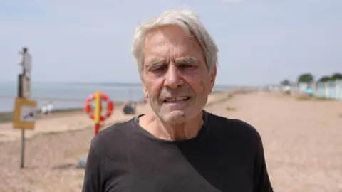Qays Najm/BBC Stephen Vince has grey hair and is wearing a black top. He is standing on the beach.