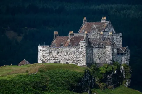 Michaela Murray A large stone castle stands on a rocky cliff surrounded by green countryside. The historic building features thick stone walls, multiple chimneys and steep roofs, with a dense forest forming a dark backdrop.